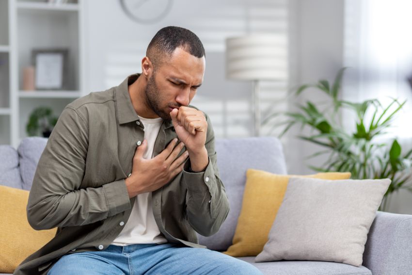 Junger afroamerikanischer Mann sitzt zu Hause auf der Couch und hustet. Leidet unter einem Asthmaanfall und Allergien. Er hält sich die Brust, bedeckt seinen Mund mit der Hand